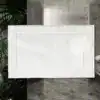 Biały dywanik łazienkowy z ozdobnym obramowaniem, na tle betonowej ściany i rośliny.